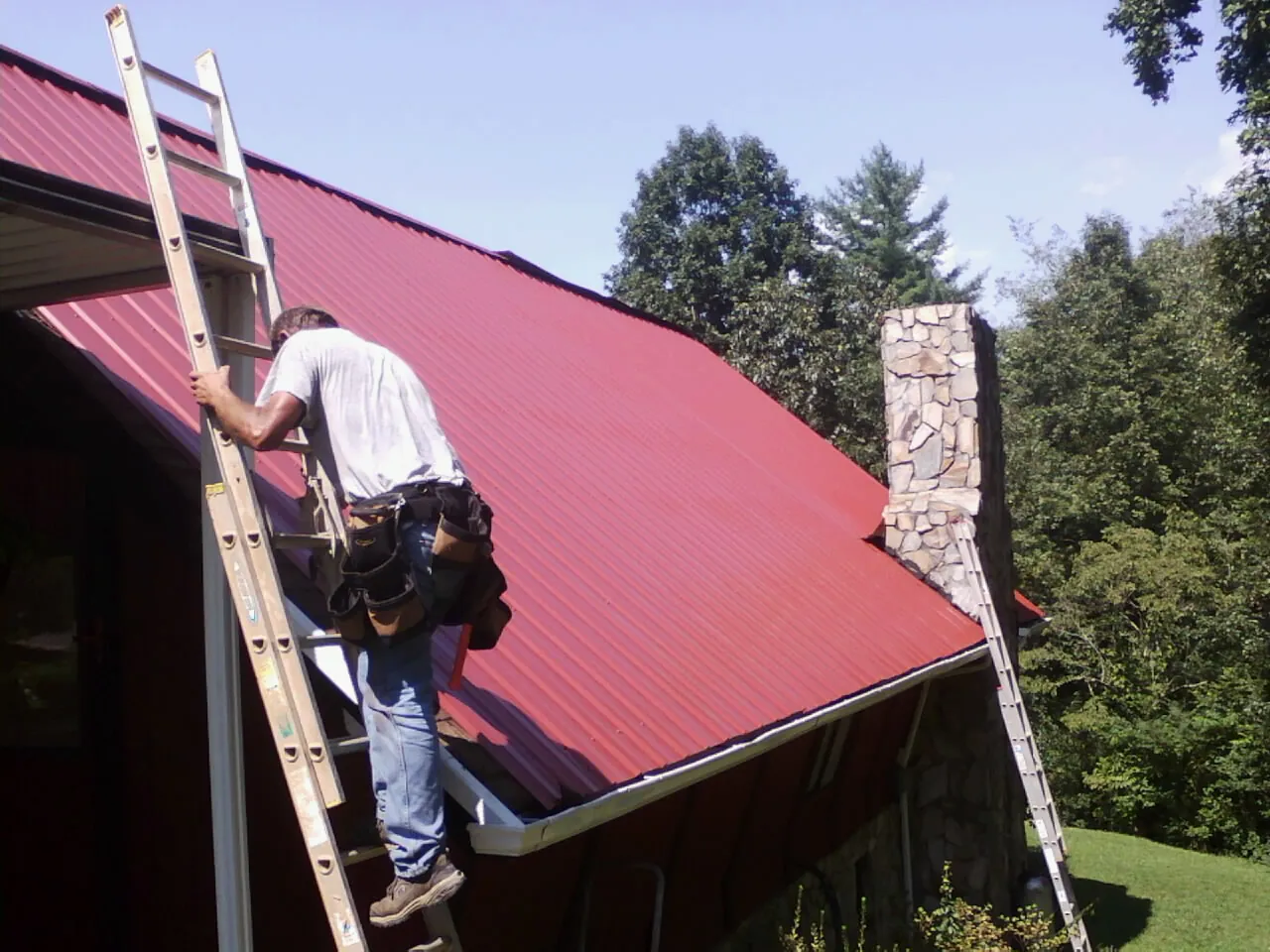 Roofer on a ladder beside a red metal roof and stone chimney — Soffit & Fascia Repair by Roofing Brothers in Rogersville